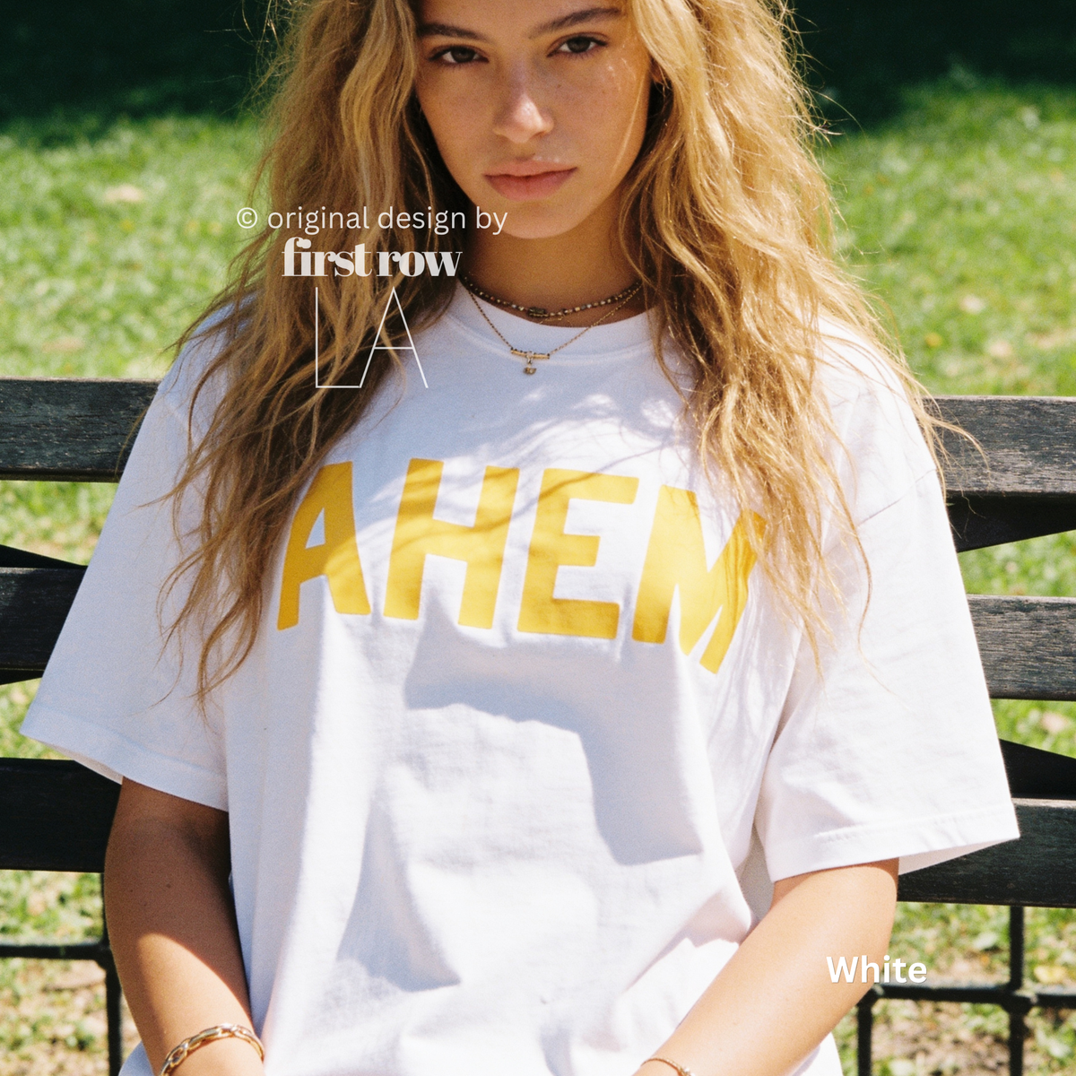A woman sitting on a wooden bench outdoors wearing an oversized white graphic T-shirt with the word 'AHEM' printed in bold yellow letters across the chest. 

Original design and copyright by first row LA, August 4th 2026.