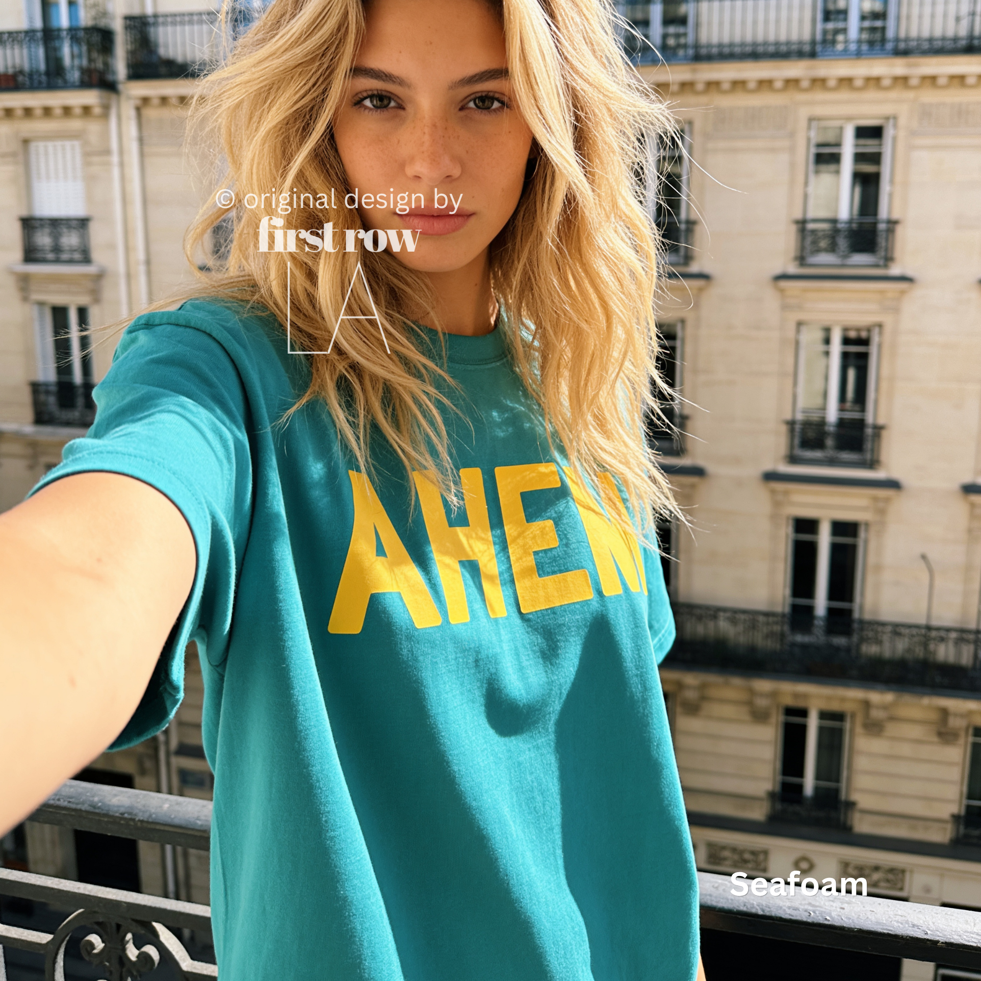 A woman taking a selfie outdoors on a balcony, wearing a teal t-shirt with large yellow text reading 'AHEM'. 

Original design and copyright by first row LA, August 4th 2026.