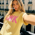 A woman taking a selfie outdoors wearing a pale yellow oversized T-shirt with large pink 'AWK' lettering across the chest. 

Original design and copyright by first row LA, August 4th 2026.