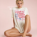 A woman sits against a soft pink studio background wearing an oversized cream T-shirt with a colorful graphic print that reads, 'A YAWN IS A SILENT SCREAM FOR COFFEE, Tired Peoples CoffeeClub.' Original design by firstrow LA.