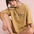 A woman wearing a mustard yellow oversized T-shirt with the black printed text 'BE KIND of a badass' on the front, posed against a soft pink studio background. White text overlay reads 'original design by first row LA'.