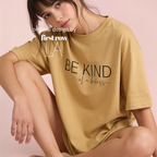 A woman wearing a mustard yellow oversized T-shirt with the black printed text 'BE KIND of a badass' on the front, posed against a soft pink studio background. White text overlay reads 'original design by first row LA'.
