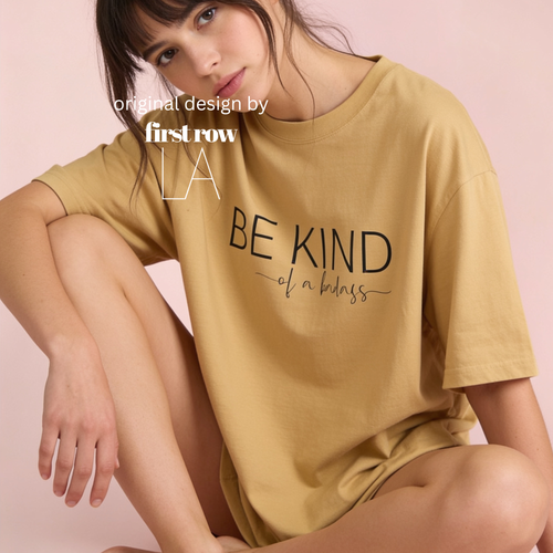 A woman wearing a mustard yellow oversized T-shirt with the black printed text 'BE KIND of a badass' on the front, posed against a soft pink studio background. White text overlay reads 'original design by first row LA'.