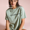 A woman wearing a light sage green oversized t-shirt with the black printed text 'C'EST LA FUCKING VIE' across the chest, posed against a solid pale pink background.