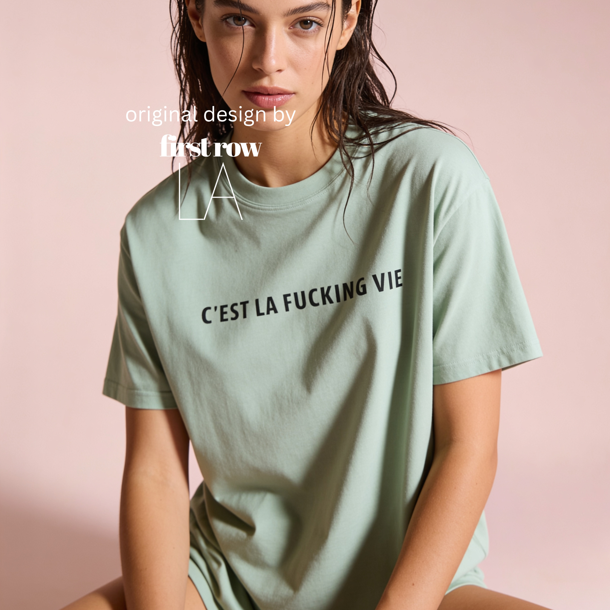 A woman wearing a light sage green oversized t-shirt with the black printed text 'C'EST LA FUCKING VIE' across the chest, posed against a solid pale pink background.