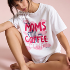 A woman wearing a white graphic t-shirt with pink and blue text reading 'Lucky Moms Coffee Club, All Day, Every Day' and a coffee themed illustrations on a background of pink clouds. Original design by first row LA.