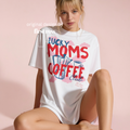 A woman wearing an oversized white graphic T-shirt with the words 'LUCKY MOMS COFFEE Club, All Day, Every Day' in red and blue, on a background of pink clouds. Original design by first row LA.