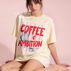 A woman wearing an oversized cream-colored graphic t-shirt with red lettering that reads 'COFFEE & AMBITION, on rereat' and a blue coffee pot illustration on a pink striped background. 