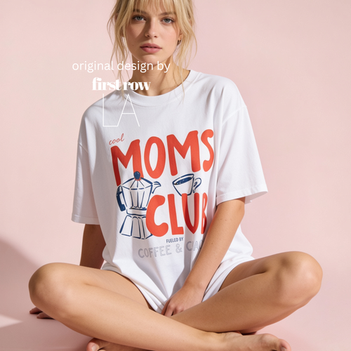 A woman sits cross-legged against a soft pink studio background wearing an oversized white graphic T-shirt with large red 'Cool MOMS CLUB, Fueled by Coffee and Chaos' lettering and coffee-themed illustrations. Designed by firstrow LA.
