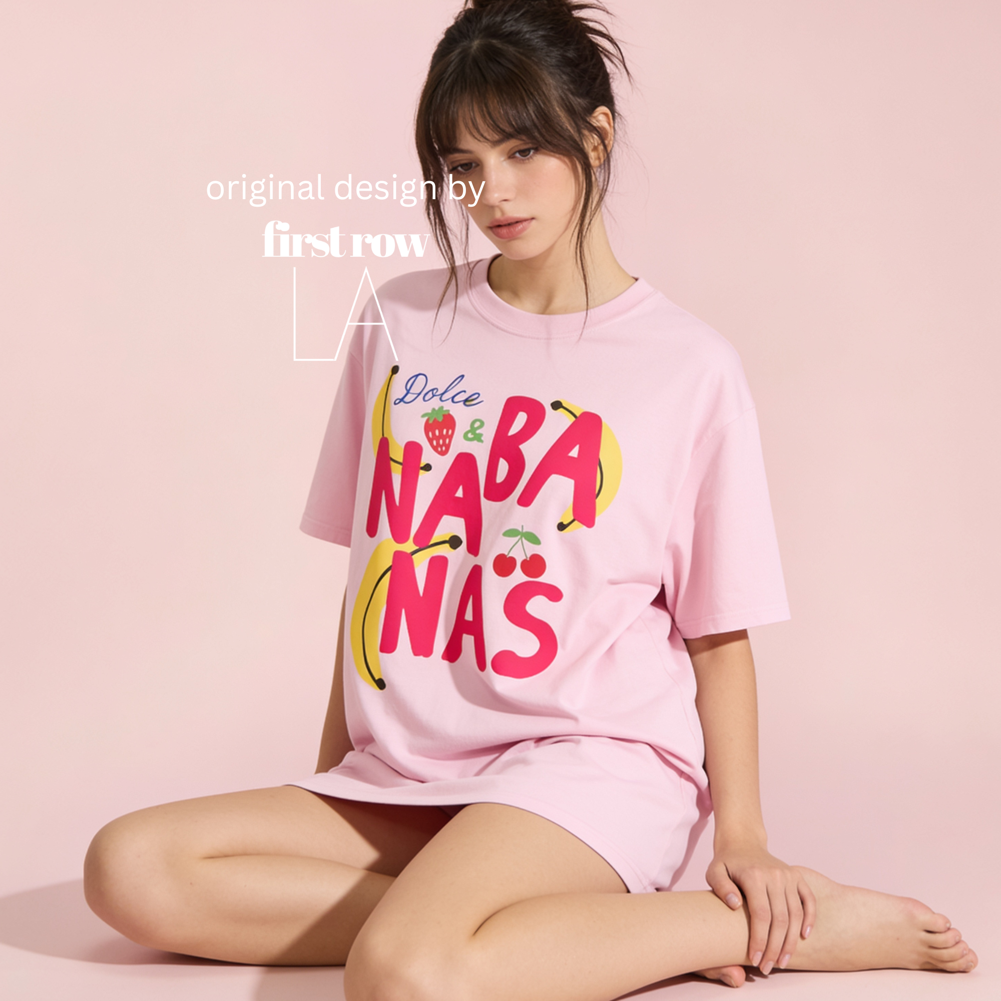 A woman sits cross-legged against a solid pale pink studio background, wearing a light pink oversized T-shirt and matching shorts. The T-shirt has a large colorful graphic reading 'Dolce and Bananas N' with banana, cherry and strawberry illustrations.