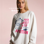 A woman wearing a sweatshirt with a graphic print that reads 'pour yourself a cup of ambition' in pink and blue, alongside a coffee pot and cup illustration. Original design by first row LA.