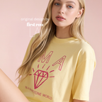 A blonde woman wearing a pale yellow graphic T-shirt with a red outlined diamond and the words 'I'm a diamond in a rhinestone world'. Original design by first row LA.