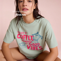 A woman wearing a light sage green graphic t-shirt with pink text that reads 'FUELED BY COFFEE POWERED BY GOOD VIBES' along with pink coffee-themed illustrations, posed against a solid pink background.