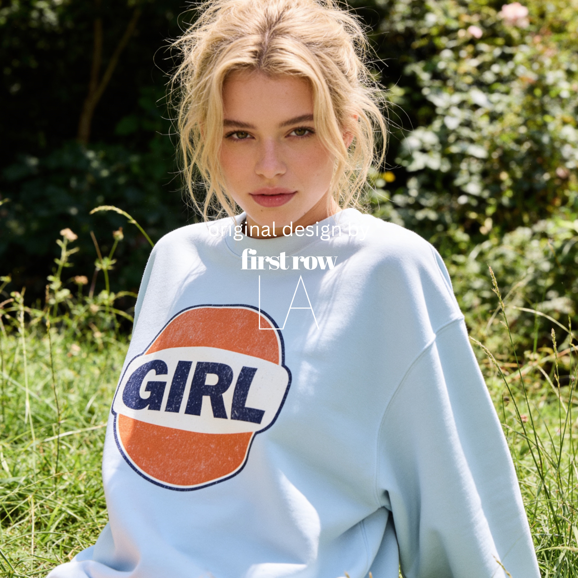 A woman outdoors wearing a light blue sweatshirt with a large retro-style orange, white, and navy 'GIRL' graphic on the front. The image has a watermark overlay reading 'Original designs first row LA'.