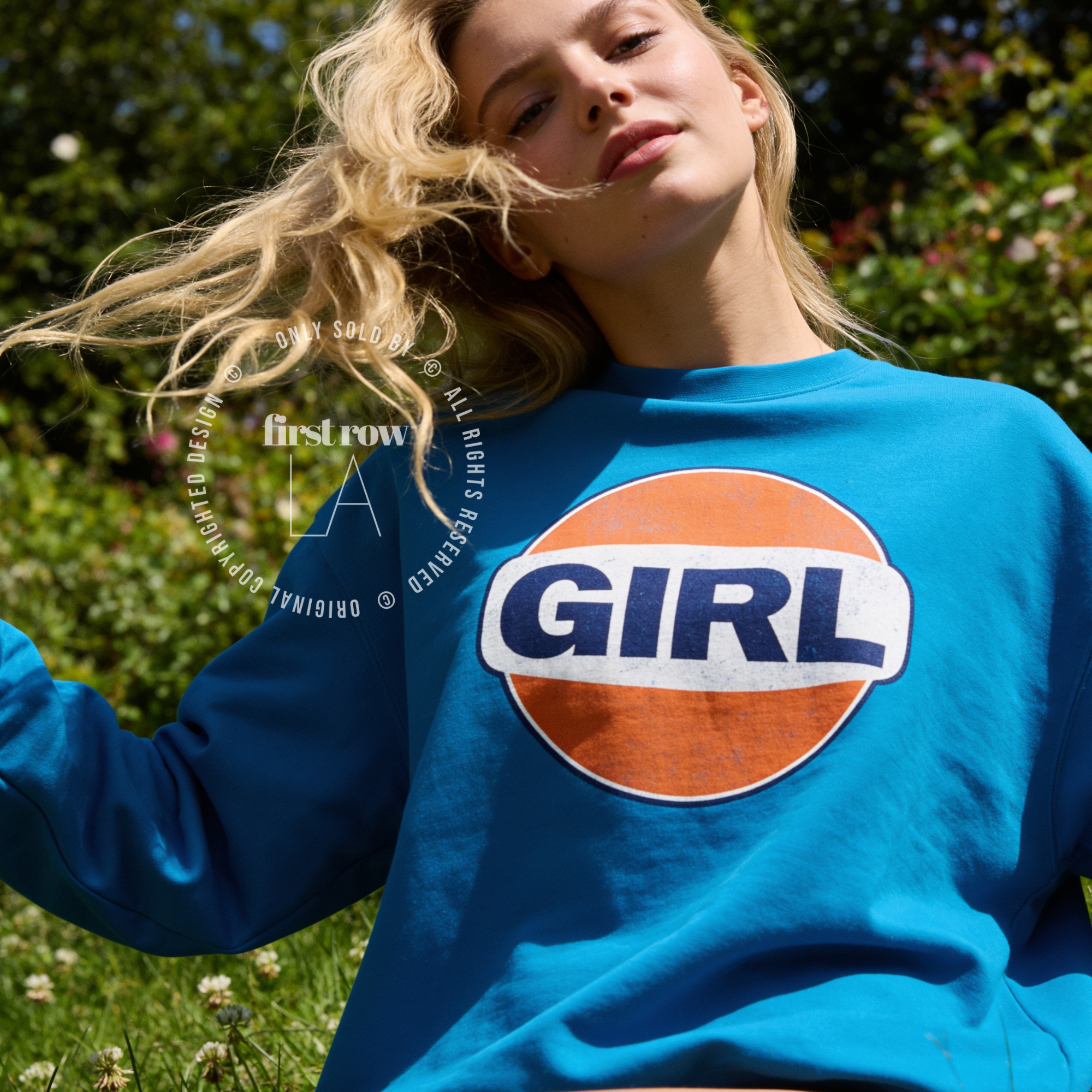 A woman outdoors wearing a bright blue sweatshirt with a large circular graphic on the chest that reads “GIRL” in navy blue letters on a white band over an orange circle.