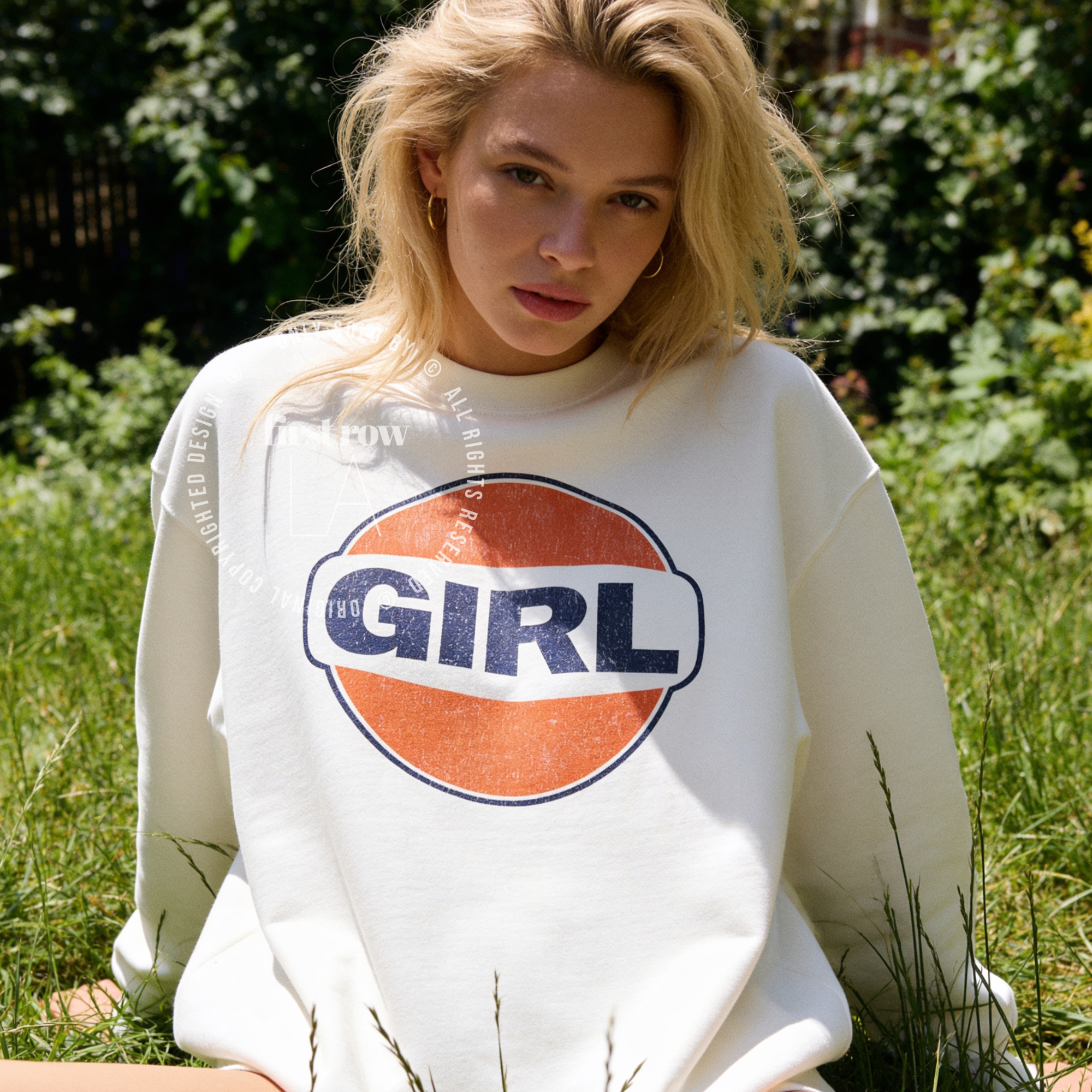 A woman outdoors in a grassy area wearing a white crewneck sweatshirt with a large retro-style circular graphic on the chest featuring the word 'GIRL' in navy blue over an orange circle with a white band.