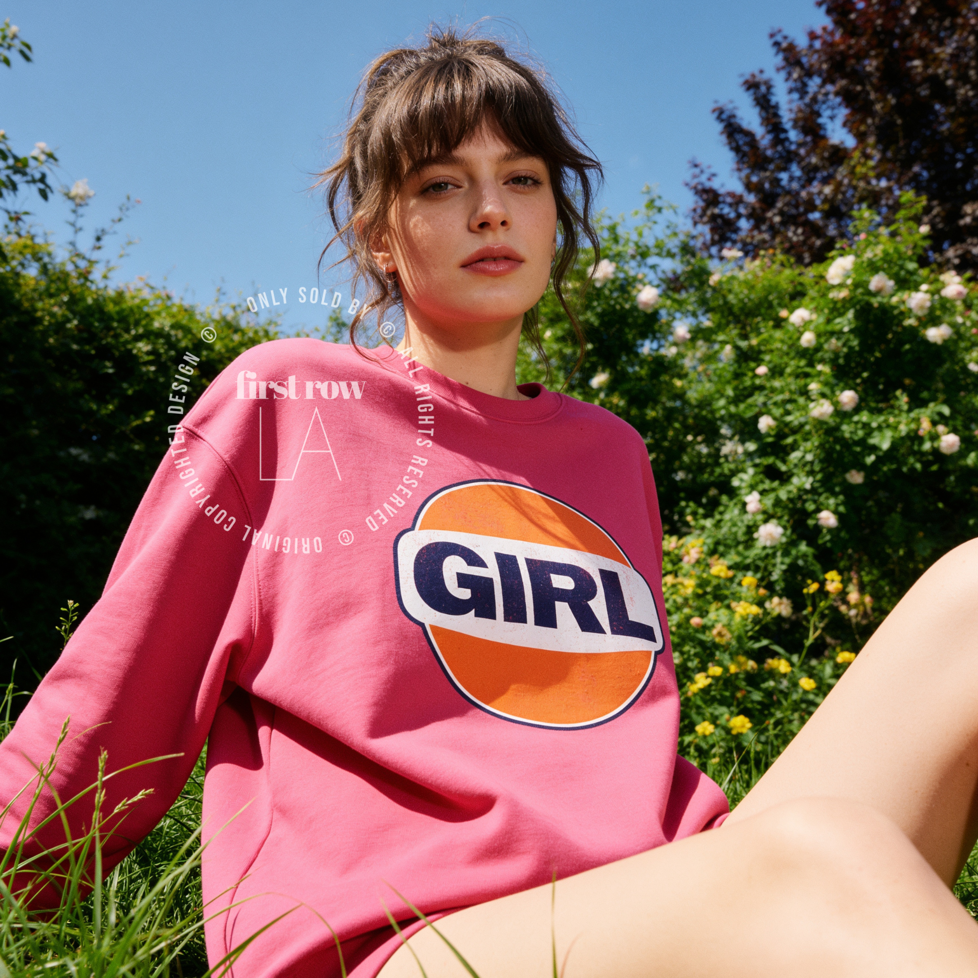 A woman outdoors wearing a pink sweatshirt with a large retro-style orange, white, and navy 'GIRL' graphic on the front. The image has a watermark overlay reading 'Original designs first row LA'.
