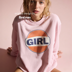 A woman wearing a light pink sweatshirt with a retro-style circular graphic on the chest featuring an orange circle, a white banner, and the word 'GIRL' in navy blue. Original design by first row LA.