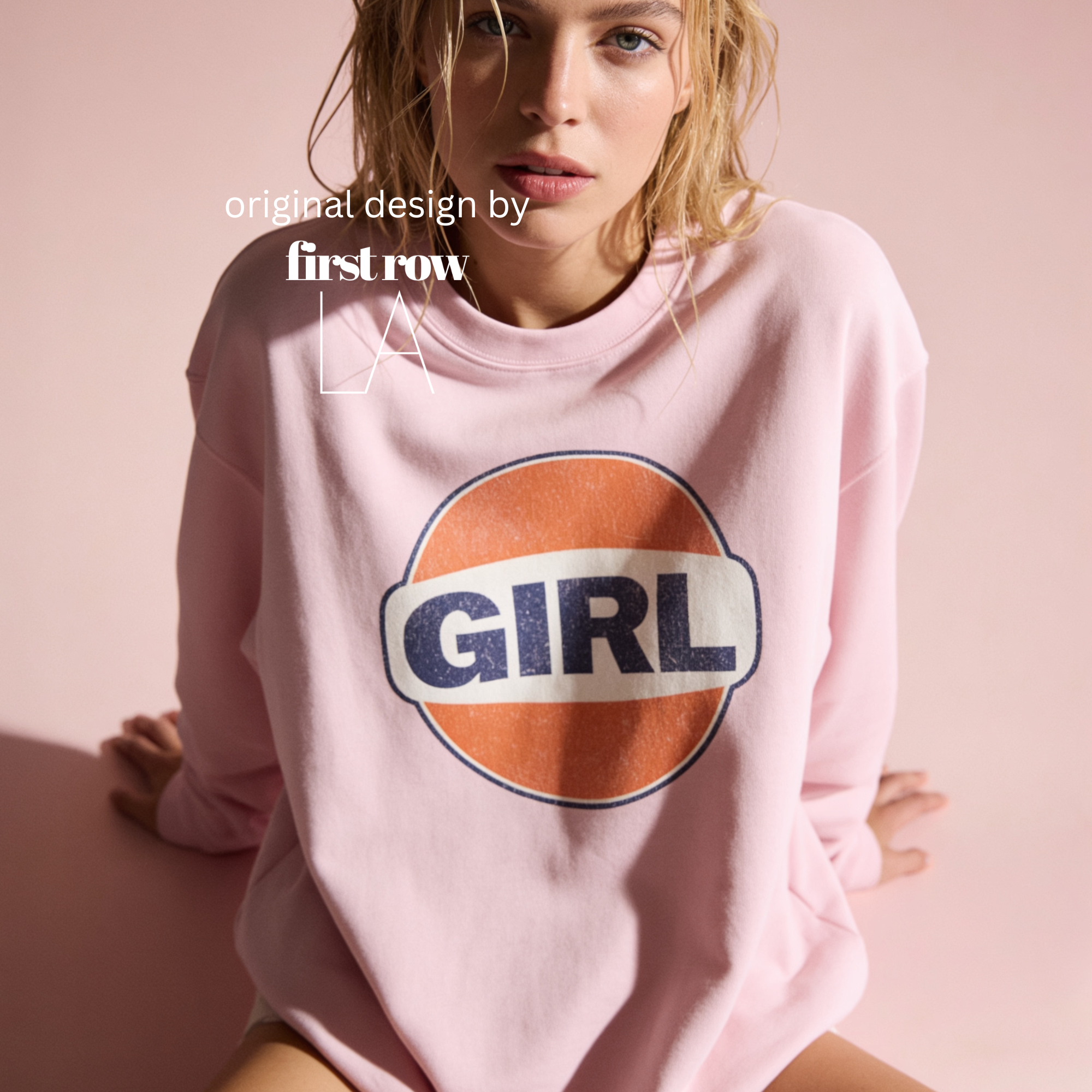 A woman wearing a light pink sweatshirt with a retro-style circular graphic on the chest featuring an orange circle, a white banner, and the word 'GIRL' in navy blue. Original design by first row LA.
