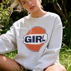 A woman outdoors wearing a ash grey sweatshirt with a large retro-style orange, white, and navy 'GIRL' graphic on the front. The image has a watermark overlay reading 'Original designs first row LA'.