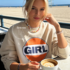 A woman sitting at a beach-side table wearing a cream sweatshirt with a large orange, white, and navy 'GIRL' graphic on the front. She is holding a cup of coffee, with a beach and ocean in the background.