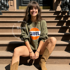A woman sits on the stone steps of a brownstone building, wearing an olive green sweatshirt with a large orange and white circular 'GIRL' graphic on the front, paired with tan cowboy boots.