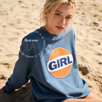 A woman outdoors wearing a blue sweatshirt with a large retro-style orange, white, and navy 'GIRL' graphic on the front. The image has a watermark overlay reading 'Original designs first row LA'.