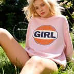 A woman sitting outdoors on grass wearing a light pink sweatshirt with a large retro-style circular graphic on the chest that reads “GIRL” in dark blue letters inside an orange circle with a white center.