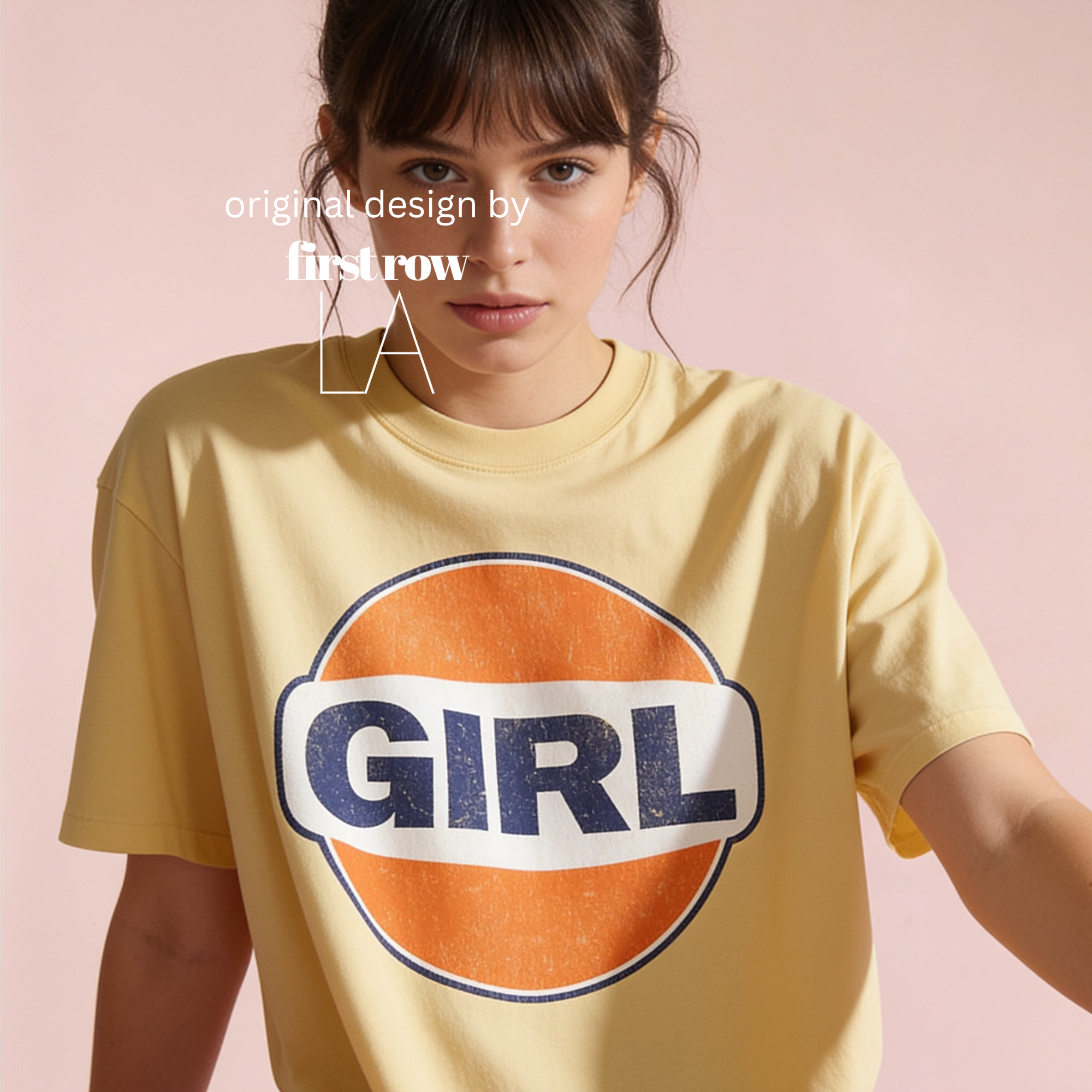 A woman wearing a pale yellow graphic T-shirt with a large orange, white, and navy circular 'GIRL' logo on the front, photographed against a soft pink studio background. Designed by first row LA.