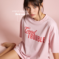 A woman wearing a light pink oversized T-shirt with 'Good TROUBLE' printed in red across the chest, posed against a soft pink background. Design by first row LA.