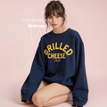 A woman sits against a solid pale pink background, wearing a navy blue crewneck sweatshirt with 'GRILLED CHEESE Club' printed in yellow on the front. Original design by first row LA.