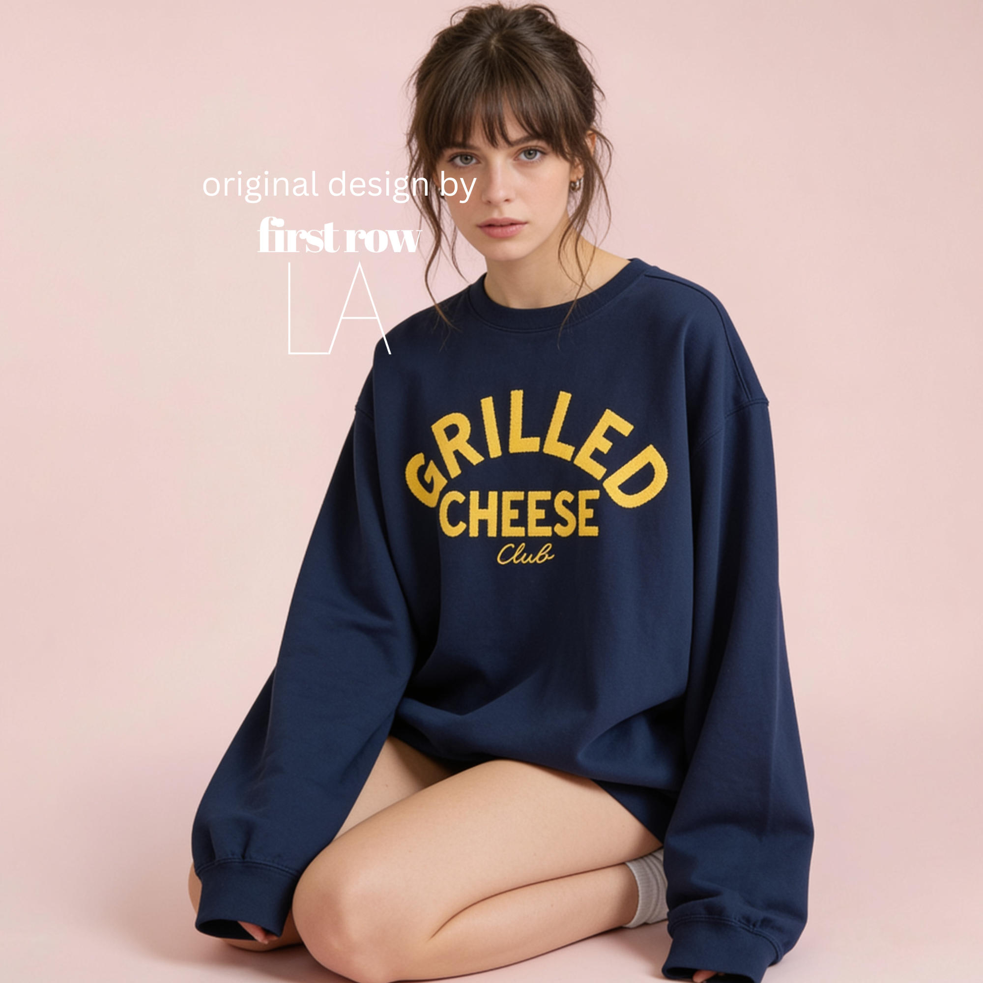 A woman sits against a solid pale pink background, wearing a navy blue crewneck sweatshirt with 'GRILLED CHEESE Club' printed in yellow on the front. Original design by first row LA.