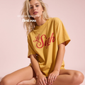A woman sitting cross-legged wearing an oversized mustard yellow t-shirt with 'Le Sigh' printed in red cursive across the chest, set against a solid pale pink background.