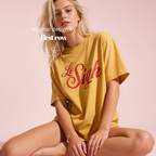 A woman sitting cross-legged wearing an oversized mustard yellow t-shirt with 'Le Sigh' printed in red cursive across the chest, set against a solid pale pink background.