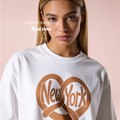 A woman wearing a white crewneck sweatshirt with a brown graphic of a pretzel forming a heart shape around the words 'New York' in script. Original design by first row LA.