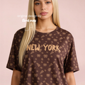 A blonde model wearing a brown oversized t-shirt with a golden pretzel and pizza pattern and 'NEW YORK' printed across the chest, set against a solid pale pink background. Original design by first row LA.