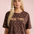 A blonde model wearing a brown oversized t-shirt with a golden pretzel and pizza pattern and 'NEW YORK' printed across the chest, set against a solid pale pink background. Original design by first row LA.