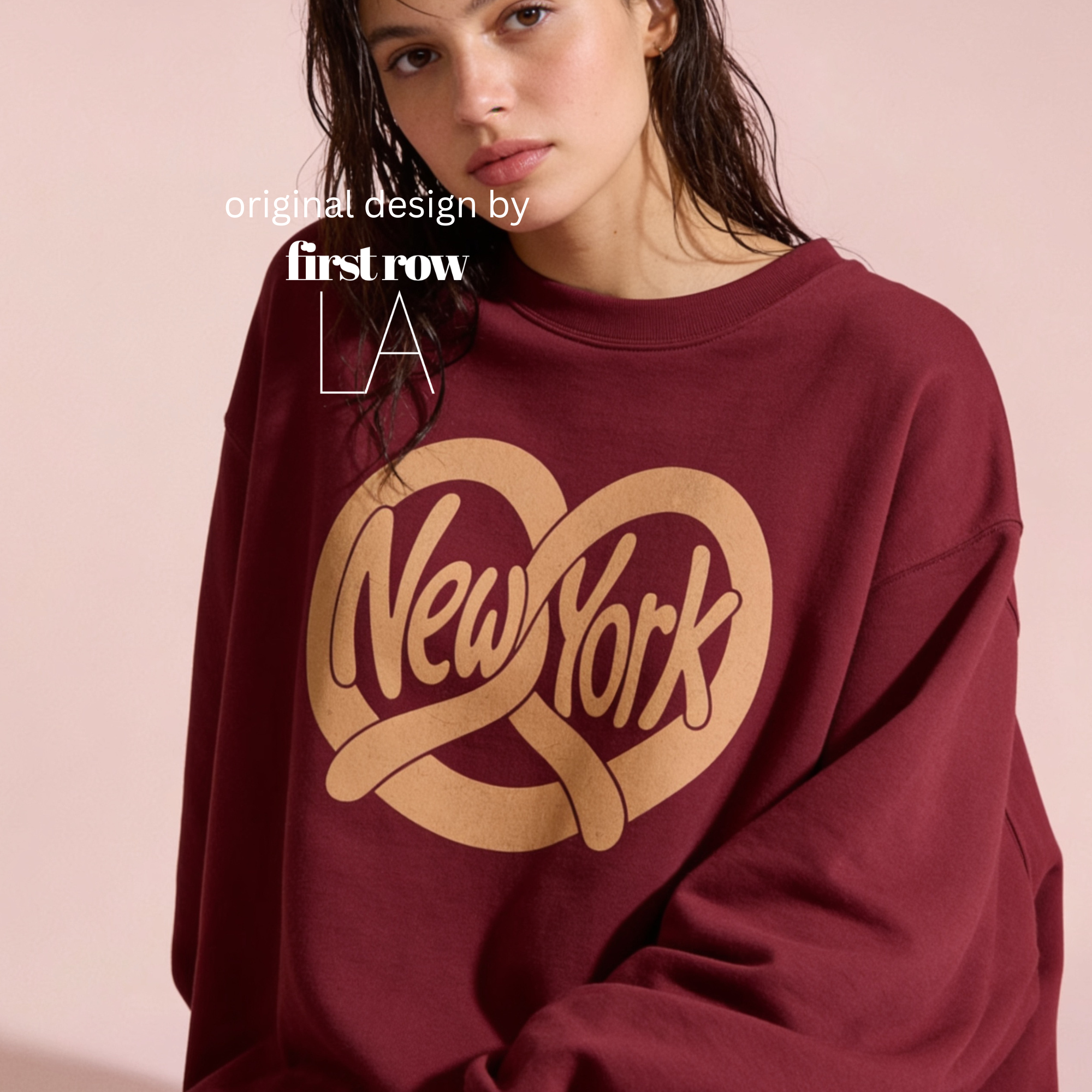 A woman wearing an oversized maroon sweatshirt with a large tan 'New York' pretzel-heart graphic on the front. Original design by first row LA'.
