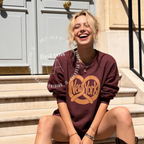 A woman sitting on stone steps wearing a heather maroon sweatshirt with a large tan 'New York' pretzel graphic on the front. Original design by first row LA.