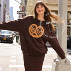A woman stands on a city street wearing a dark brown sweatshirt and matching pants set. The sweatshirt features a large tan 'New York' graphic. Original design by first row LA.