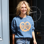 A woman wearing a blue crewneck sweatshirt with a large yellow 'New York' pretzel graphic, paired with blue jeans, standing in front of a dark doorway. Original design by first row LA.