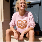 A woman sitting outdoors in front of a dark blue door, wearing a light pink crewneck sweatshirt with a large brown pretzel New York graphic on the front. Original design by first row LA.