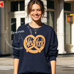 A woman wearing a navy blue sweatshirt with a large tan pretzel-shaped graphic and the words 'New York' across the front, standing outdoors in front of a building. Original design by first row LA.