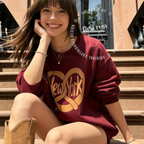 A woman sitting on outdoor stone steps wearing an oversized burgundy sweatshirt with a large gold 'New York' pretzel graphic. Original design by first row LA.