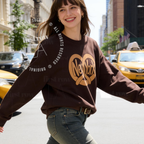 A woman walking on a city street wearing a dark brown sweatshirt with a tan New York heart graphic and blue jeans. Original design by first row LA.