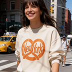 A woman smiling on a city street wearing a cream sweatshirt with a large orange pretzel graphic and the words 'New York' across the front. Original design by first row LA.