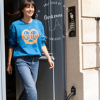 A woman wearing a bright blue crewneck sweatshirt with a yellow and orange New York City skyline graphic, paired with blue jeans, walking out of a building. Original design by first row LA.