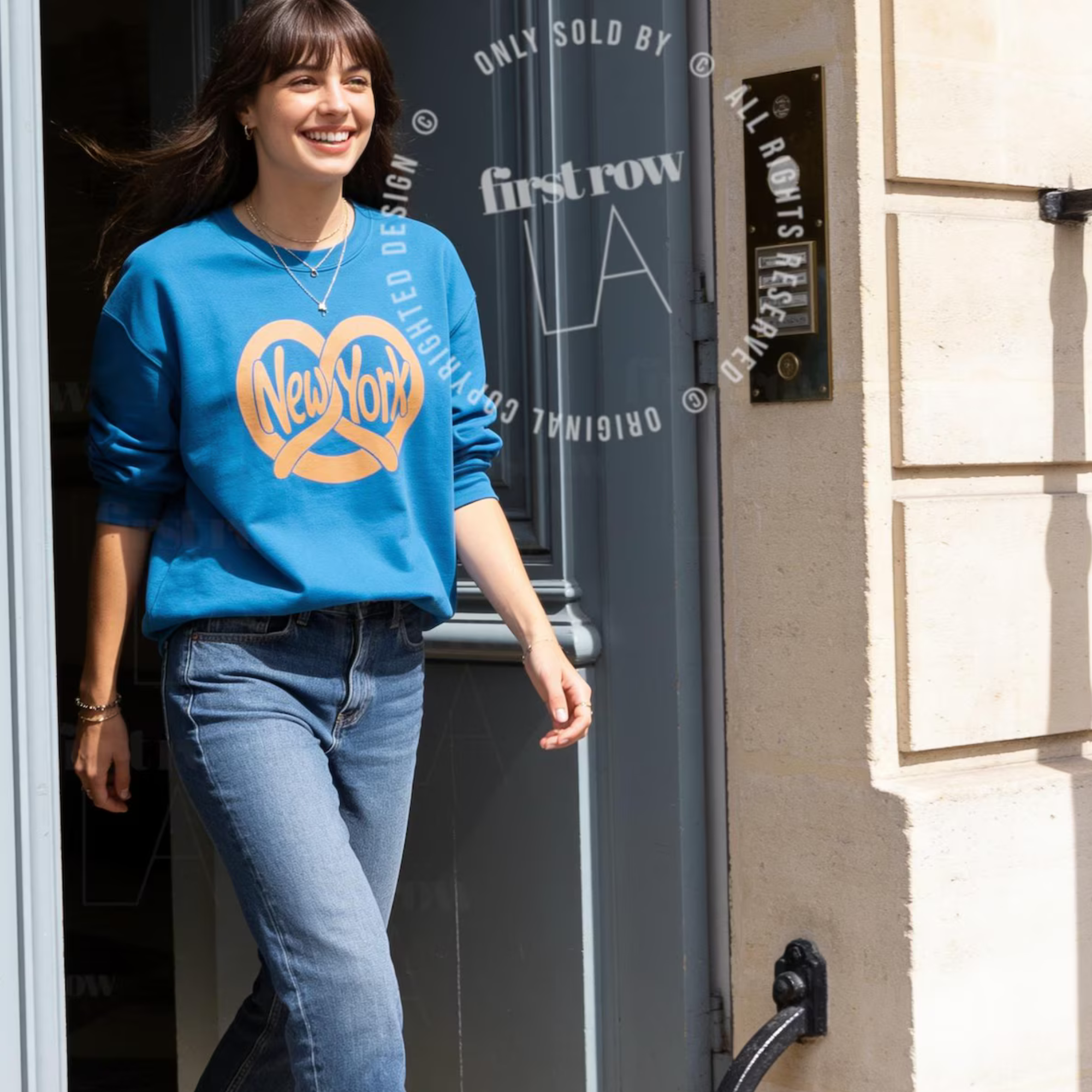 A woman wearing a bright blue crewneck sweatshirt with a yellow and orange New York City skyline graphic, paired with blue jeans, walking out of a building. Original design by first row LA.