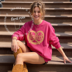 A woman sitting on outdoor stone steps wearing a bright pink crewneck sweatshirt with a large gold heart and pretzel graphic that says 'New York'. Original design by first row LA.