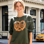 A woman wearing an olive green crewneck sweatshirt with a large orange 'New York' heart and pretzel graphic on the front, standing outdoors in front of a city building. Original design by first row LA.
